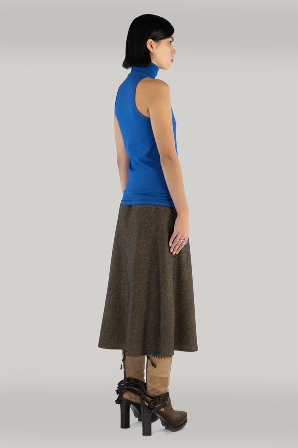A woman standing in front of grey backdrop wearing a cobalt blue sleeveless turtleneck, paired with a brown long wool skirt. She is also wearing tall brown heeled boots.